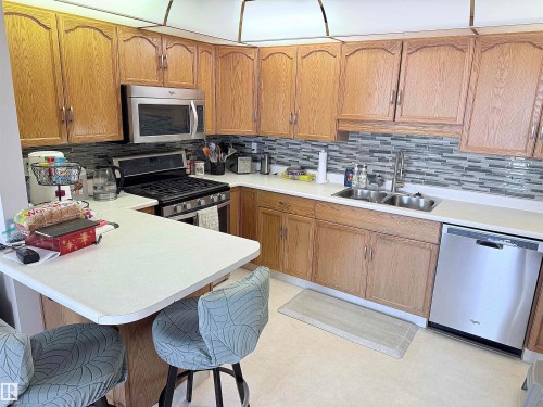 Kitchen featuring wood cabinetry, white countertops, stainless steel appliances, a tile backsplash, and a breakfast bar - 410 9926 100 Avenue, Fort Saskatchewan, AB - Indoor Photo Showing Kitchen With Double Sink