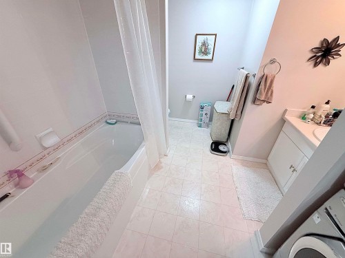 Bathroom featuring a white bathtub with a shower curtain, white tile flooring, a single vanity with a white countertop, and a toilet - 410 9926 100 Avenue, Fort Saskatchewan, AB - Indoor Photo Showing Bathroom
