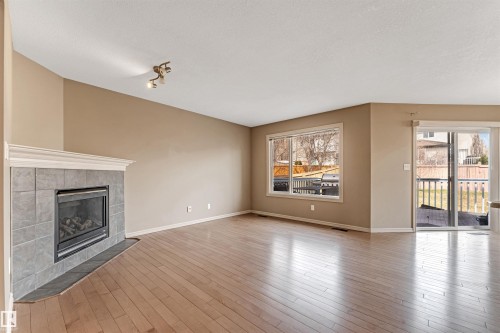 Living area featuring a corner fireplace with a tiled surround and white mantel, wood-finish flooring, a large window, and a sliding glass door leading to an outdoor deck - 1431 Hodgson Way, Edmonton, AB - Indoor Photo Showing Living Room With Fireplace