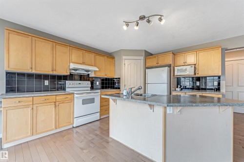 Kitchen featuring wood-finish cabinetry, black tile backsplash, stainless steel sink, a functional island, and track lighting - 1431 Hodgson Way, Edmonton, AB - Indoor Photo Showing Kitchen With Double Sink