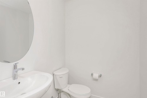 White-walled washroom featuring a circular mirror, a wall-mounted sink with a chrome faucet, and a white toilet - 9552 Carson Bend Bend, Edmonton, AB - Indoor Photo Showing Bathroom