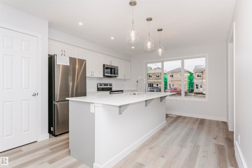 Modern kitchen featuring a central island with a sink, white cabinetry, stainless steel appliances, pendant lighting, and wood-finish flooring - 9564 Carson Bend Bend, Edmonton, AB - Indoor Photo Showing Kitchen With Upgraded Kitchen