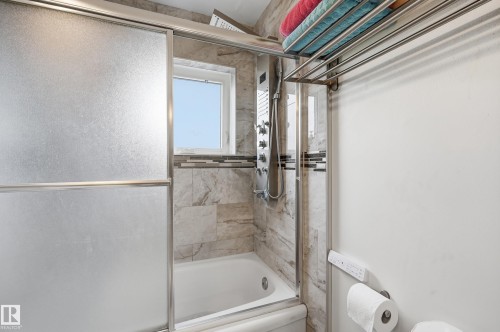 Bathtub and shower combination featuring a multi-setting shower panel, frosted sliding glass doors, a window, and natural stone-look wall tiling with a decorative mosaic accent - 105 7204 81 Avenue, Edmonton, AB - Indoor Photo Showing Bathroom