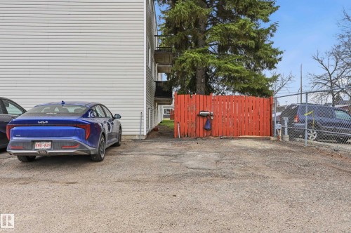 Paved parking area featuring a red slatted fence and mature evergreen trees - 105 7204 81 Avenue, Edmonton, AB - Outdoor
