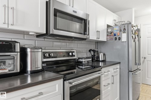 Kitchen featuring white shaker-style cabinetry with brushed nickel hardware, stainless steel appliances, dark countertops, and a light gray subway tile backsplash - 105 7204 81 Avenue, Edmonton, AB - Indoor Photo Showing Kitchen