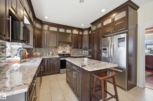 Kitchen featuring dark wood cabinetry with upper frosted glass panels, stainless steel appliances, speckled stone countertops, and a mosaic tile backsplash - 3105 65 Street, Beaumont, AB - Indoor Photo Showing Kitchen With Upgraded Kitchen