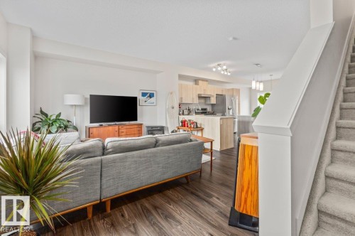 Open-concept living space featuring wood-finish flooring, a modern kitchen with light wood cabinetry and stainless steel appliances, and a carpeted staircase - 21 979 Crystallina Nera Way, Edmonton, AB - Indoor Photo Showing Living Room