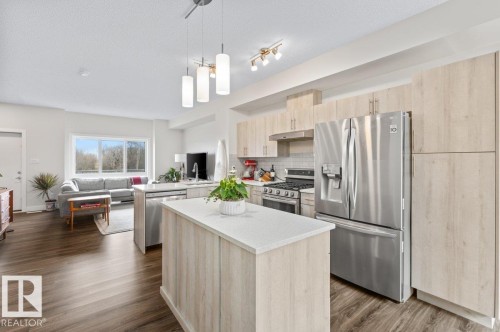 Modern open-concept kitchen featuring light wood-finish cabinetry, stainless steel appliances, a center island with a white countertop, and wood-finish flooring - 21 979 Crystallina Nera Way, Edmonton, AB - Indoor Photo Showing Kitchen