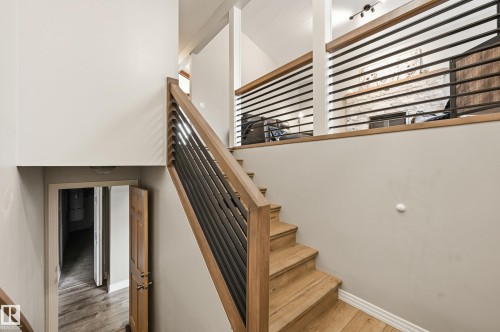 Hardwood staircase with modern metal railing, wood-finish flooring, white painted walls, and an upper-level loft area featuring a white brick fireplace with a dark mantel - 1729 65 Street, Edmonton, AB - Indoor Photo Showing Other Room