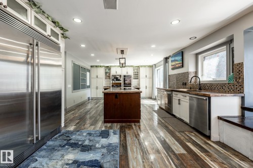 Gourmet kitchen featuring a large central island with a dark countertop, extensive wood-finish flooring, stainless steel appliances, white cabinetry, and a mosaic tile backsplash - 7 Doolan Place, St. Albert, AB - Indoor Photo Showing Kitchen With Upgraded Kitchen