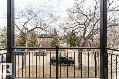 Balcony with black metal railings and structural supports, offering views of a tree-lined street and brick residences - 204 10126 144 Street, Edmonton, AB - Outdoor With Balcony