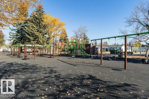 Community playground featuring expansive rubberized surfacing, multiple play structures with green and brown accents, swing sets, and mature perimeter trees - 204 10126 144 Street, Edmonton, AB - Outdoor