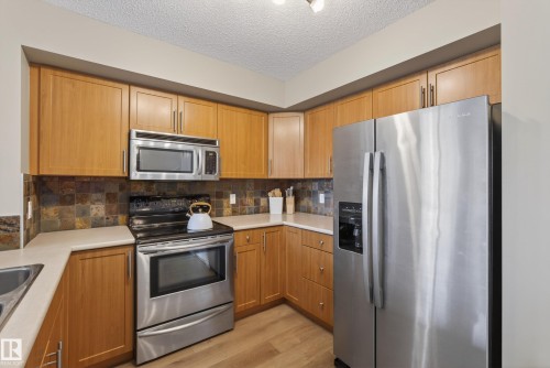 Kitchen featuring wood cabinetry, stainless steel appliances, light-toned countertops, a mosaic tile backsplash, and wood-finish flooring - 17 171 Brintnell Boulevard, Edmonton, AB - Indoor Photo Showing Kitchen With Stainless Steel Kitchen