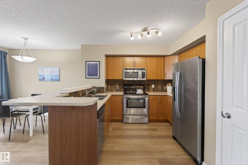 Kitchen featuring wood-finish flooring, stainless steel appliances, wood cabinetry, a tile backsplash, and an integrated kitchen island with a breakfast bar - 17 171 Brintnell Boulevard, Edmonton, AB - Indoor Photo Showing Kitchen With Stainless Steel Kitchen With Double Sink