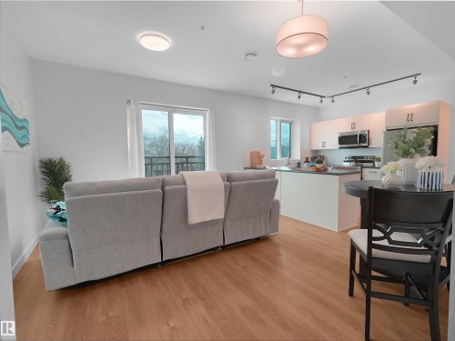 Open-concept living area featuring wood-finish flooring, a contemporary kitchen with white cabinetry and stainless steel appliances, a kitchen island with seating, a sliding glass door leading to a balcony, and a modern overhead light fixture - 403 10115 88 Avenue, Edmonton, AB - Indoor