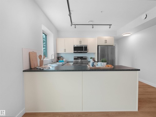 Kitchen featuring a peninsula island with dark countertops, light cabinetry, stainless steel appliances, and wood-finish flooring - 403 10115 88 Avenue, Edmonton, AB - Indoor Photo Showing Kitchen
