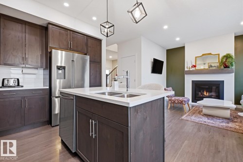 The kitchen features a stainless steel refrigerator and dishwasher, dark wood cabinetry, and a kitchen island with a double sink - 405 Genesis Court, Stony Plain, AB - Indoor Photo Showing Kitchen With Fireplace With Double Sink