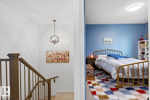 This property features a bedroom with blue walls and a bed frame with a metallic finish, visible from a hallway with a staircase and a decorative light fixture - 405 Genesis Court, Stony Plain, AB - Indoor
