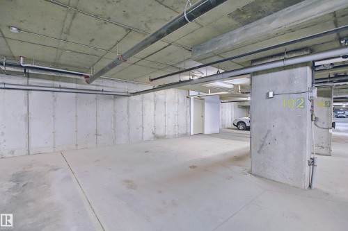 Concrete parking space featuring exposed concrete walls and ceiling, overhead piping, and fluorescent lighting - 114 3207 James Mowatt Trail, Edmonton, AB - Indoor Photo Showing Garage