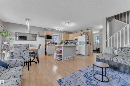 Open concept living area featuring wood-finish flooring throughout, a kitchen with light wood cabinetry and a central island, and a dining area with a contemporary linear light fixture - 64 4350 23 Street, Edmonton, AB - Indoor
