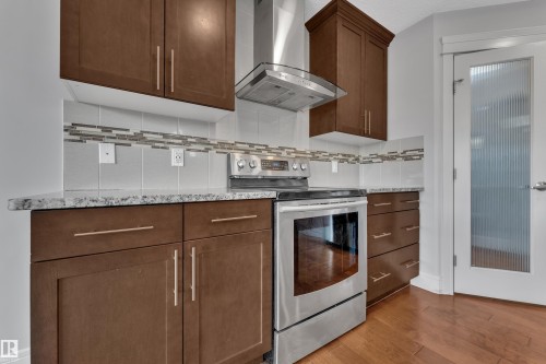 Kitchen featuring stainless steel appliances, dark wood-finish cabinetry, granite-patterned countertops, a tile backsplash with mosaic accents, and wood-finish flooring - 3363 13 Avenue, Edmonton, AB - Indoor Photo Showing Kitchen