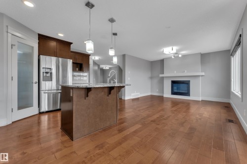 Open-concept living space with wood-finish flooring, a kitchen island with pendant lighting, a built-in stainless steel refrigerator, and a fireplace with a mantel - 3363 13 Avenue, Edmonton, AB - Indoor Photo Showing Kitchen With Upgraded Kitchen