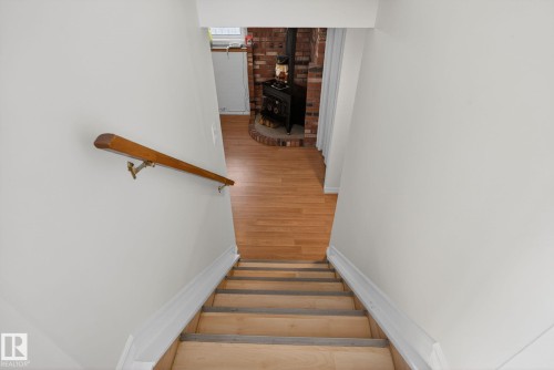 Staircase with a wood handrail descending to a lower level featuring wood-finish flooring, a brick accent wall, and a black wood-burning stove with a brick hearth - 8403 190 Street, Edmonton, AB - Indoor Photo Showing Other Room