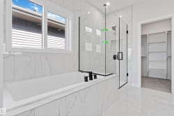 Full bathroom featuring a rectangular soaking tub, glass-enclosed shower with black fixtures, white marble-look tile flooring, and a walk-in closet with built-in shelving - 