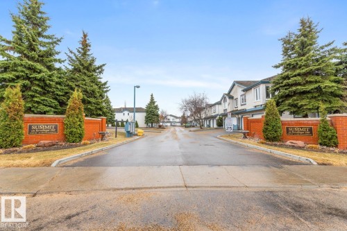 Community entrance featuring brick signage with metal lettering, mature evergreen trees, and manicured shrubs - 47 1130 Falconer Road, Edmonton, AB - Outdoor