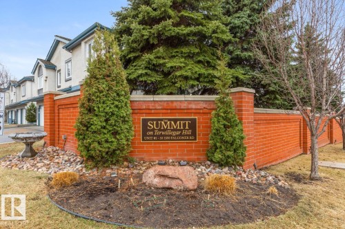Brick wall entrance with a "Summit on Terwillegar Hill" sign, featuring decorative landscaping with evergreen shrubs, natural rocks, and a stone birdbath - 47 1130 Falconer Road, Edmonton, AB - Outdoor