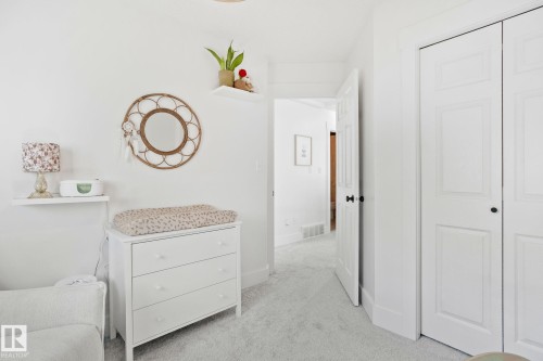 White interior with light grey carpet, featuring a white dresser, a decorative round mirror, a floating shelf, and white paneled bifold closet doors - 53 2503 24 Street, Edmonton, AB - Indoor