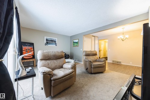 The living area features carpet flooring, neutral colored walls, and a view into an additional space with wood-look flooring and a chandelier - Sherwood Park, AB - Indoor Photo Showing Living Room