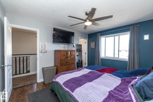 Interior room featuring a large window, a ceiling fan with integrated lighting, wood-finish flooring, and a doorway opening to a staircase with a black railing - 65 Birchmont Drive, Leduc, AB - Indoor Photo Showing Bedroom