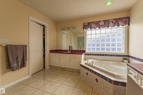 Spacious bathroom featuring a built-in soaking tub with decorative tile surround, a large glass block window, a vanity with a solid surface countertop, and white ceramic tile flooring - 195 Kulawy Drive, Edmonton, AB - Indoor Photo Showing Bathroom