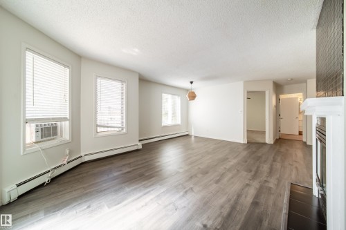 Spacious living area featuring wood-finish flooring, multiple windows with blinds, and baseboard heating - 302 9138 83 Avenue, Edmonton, AB - Indoor With Fireplace