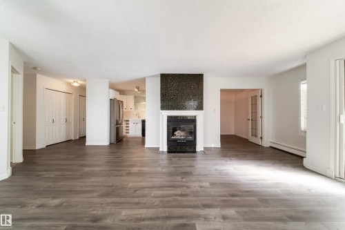 Spacious living area featuring wood-finish flooring, a fireplace with a dark mosaic tile surround and white mantel, and ample natural light from the sliding glass door - 302 9138 83 Avenue, Edmonton, AB - Indoor Photo Showing Living Room With Fireplace