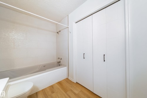 Bathroom featuring a white bathtub with a tiled shower surround, light wood-finish flooring, and white bi-fold closet doors with black hardware - 302 9138 83 Avenue, Edmonton, AB - Indoor Photo Showing Bathroom