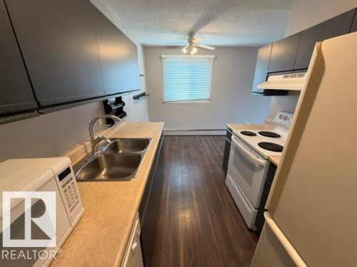 Kitchen featuring dark wood-finish cabinetry, a dual basin stainless steel sink with a gooseneck faucet, light-toned countertops, and wood-finish flooring - 201 11324 97 Street Nw, Edmonton, AB - Indoor Photo Showing Kitchen With Double Sink