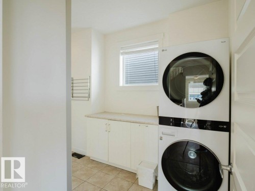 Dedicated laundry area featuring a stacked washer and dryer, tiled flooring, white cabinetry, a stone-finish countertop, and a window with blinds - 15867 11 Avenue, Edmonton, AB - Indoor Photo Showing Laundry Room