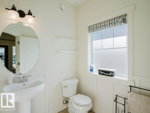 Bathroom featuring wainscoting, pedestal sink, oval mirror, and a window with a striped Roman shade - 15867 11 Avenue, Edmonton, AB - Indoor Photo Showing Bathroom