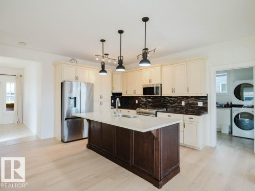 Kitchen featuring a central island with a light-toned countertop, cream-colored cabinetry, stainless steel appliances, dark backsplash, and light wood-finish flooring - 15867 11 Avenue, Edmonton, AB - Indoor Photo Showing Kitchen