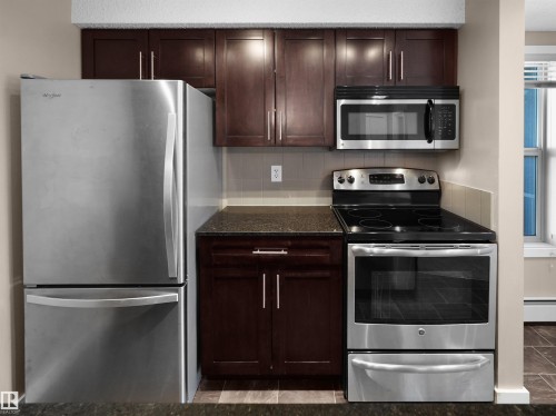 Compact kitchen featuring dark wood-finish cabinetry, stainless steel appliances, dark countertops, and a tiled backsplash - 417 1060 Mcconachie Boulevard, Edmonton, AB - Indoor Photo Showing Kitchen