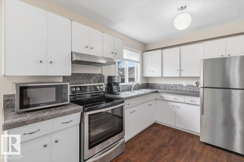 Bright kitchen featuring white cabinetry, stainless steel appliances, dark wood-finish flooring, tile countertops, and a tile backsplash - 13107 133 Street, Edmonton, AB - Indoor Photo Showing Kitchen