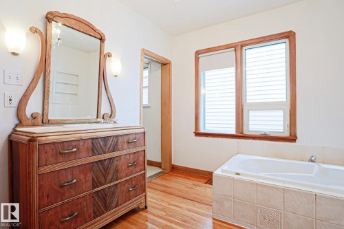 Bright bathroom featuring a built-in soaking tub with a tiled surround, a wooden window frame, and wood-finish flooring - 9611 81 Avenue, Edmonton, AB - Indoor Photo Showing Bathroom
