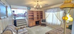 Carpeted living area featuring a built-in wood-finish shelving unit, a multi-bulb chandelier, and a large window with draped curtains - 