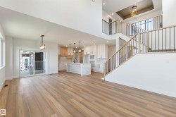 Open-concept living space featuring wood-finish flooring, a contemporary kitchen with a large island, a two-story staircase with dark metal balusters, high ceilings, and a sliding glass door opening to an exterior balcony - 