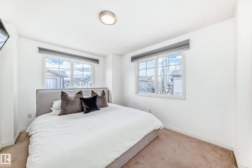 Bright bedroom featuring two large grid-pattern windows with roller shades, neutral carpeting, white walls, and a flush-mount ceiling light fixture - 45 4850 Terwillegar Common, Edmonton, AB - Indoor Photo Showing Bedroom
