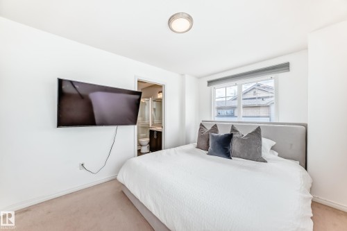 Carpeted room featuring a window with a roller shade, flush mount ceiling light, and an open doorway revealing a bathroom with a glass shower enclosure and vanity - 45 4850 Terwillegar Common, Edmonton, AB - Indoor Photo Showing Bedroom