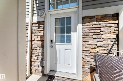 Stacked stone facade accents a white paneled door with grid glass insert, complemented by white siding and an outdoor wall sconce - 45 4850 Terwillegar Common, Edmonton, AB - Outdoor