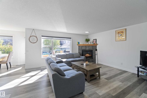 Living area with light-colored flooring, a fireplace featuring a stone surround and wooden mantel, and windows providing views of the outdoors - 1017 82 Street, Edmonton, AB - Indoor Photo Showing Living Room With Fireplace